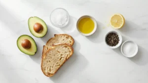 All ingredients for avocado toast laid out neatly: ripe avocado, sliced bread, lemon, salt, black pepper, and olive oil on a wooden table.