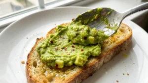 Using a spoon to spread a thick layer of chunky, seasoned avocado mash onto a warm, toasted slice of bread.