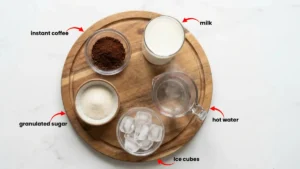 Ingredients for whipped coffee: instant coffee, sugar, hot water, milk, and ice arranged around a mixing bowl and spoon.