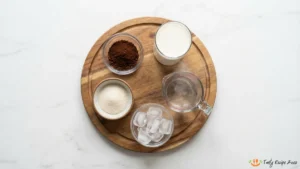 Ingredients for whipped coffee: instant coffee, sugar, hot water, milk, and ice arranged around a mixing bowl and spoon.