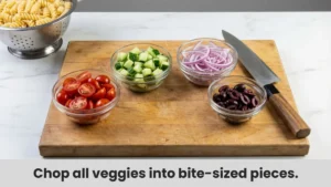 Chopping fresh cherry tomatoes, cucumbers, red onion, and olives on a cutting board for easy gluten-free pasta salad