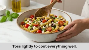 Tossing cooled gluten-free pasta with vegetables, feta cheese, and vinaigrette in a large bowl to assemble the easy pasta salad