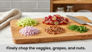 Chopped celery, red onion, grapes, pecans, and fresh herbs arranged on cutting board for chicken salad.