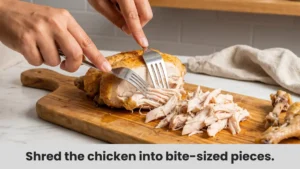 Hands using two forks to shred rotisserie chicken on wooden cutting board for chicken salad.