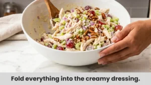 Mixing shredded chicken, grapes, celery, pecans, and onion with creamy dressing in bowl for chicken salad.