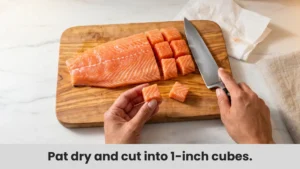 Fresh raw salmon being cut into 1-inch cubes on wooden cutting board for air fryer salmon bites recipe.