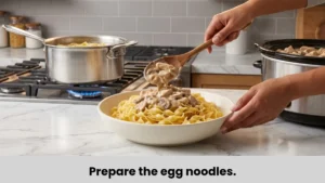 Creamy chicken stroganoff spooned over tender egg noodles beside a stainless-steel pot on the gas stove.